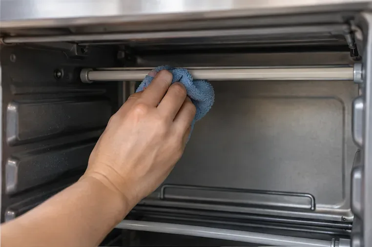 Een hand die heel voorzichtig met een zachte, vochtige microvezeldoek over een afgekoelde kwartsverwarmingsbuis in een broodrooster veegt.