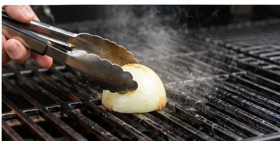 Een lange metalen tang houdt een halve ui met de snijkant naar beneden vast terwijl deze over hete grillroosters wordt gewreven, waarbij lichte stoom opstijgt.