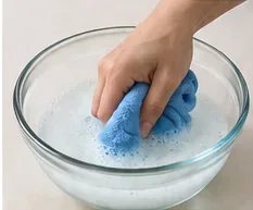 Een blauwe schoonmaakdoek wordt ondergedompeld in een glazen kom met een kleine hoeveelheid mild zeepsop.