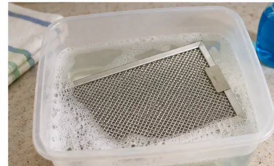 Een rechthoekig metalen vetfilter voor de magnetron, geweekt in een bak met heet zeepsop.