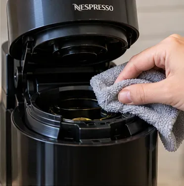 Een open Vertuo-stijl koffiezetapparaat wordt voorzichtig afgeveegd met een vochtige microvezeldoek in de buurt van het toegankelijke capsulegedeelte.