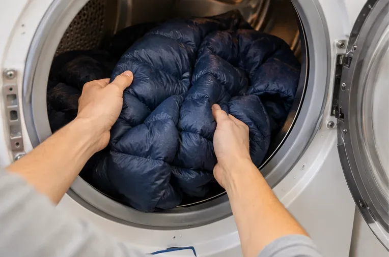 Handen maken voorzichtig de samengeklonterde isolatie in een donkerblauwe donsjas los en scheiden deze van elkaar, bij de open deur van de wasdroger.