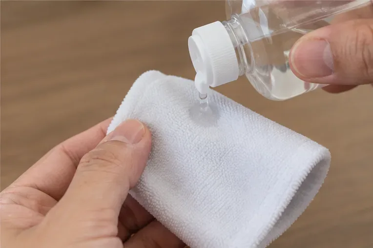 小さなボトルから透明な液体を数滴、木製の作業台の上に折りたたんだ糸くずの出ない布に垂らしている手。