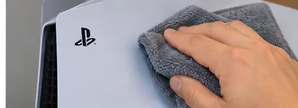 Een hand veegt het witte buitenpaneel van een PS5 af met een zachte grijze microvezeldoek.