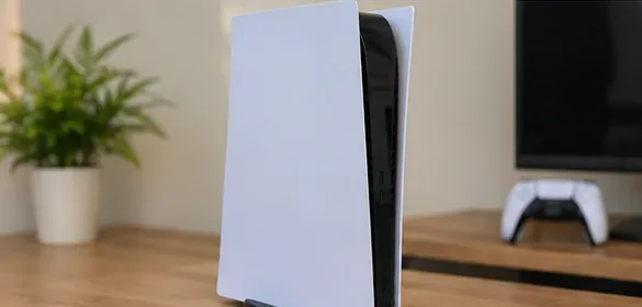 Een nette PS5 staat rechtop op een bureau met vrije ruimte eromheen en een televisie op de achtergrond.