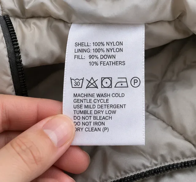 Detailopname van een wasvoorschrift op een gewatteerde jas, met symbolen voor de buitenkant, voering, vulling, wassen en drogen.