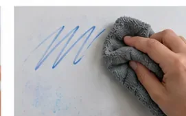 Een hand veegt een verse blauwe markerstreep weg met een zachte grijze microvezeldoek op een whiteboard.