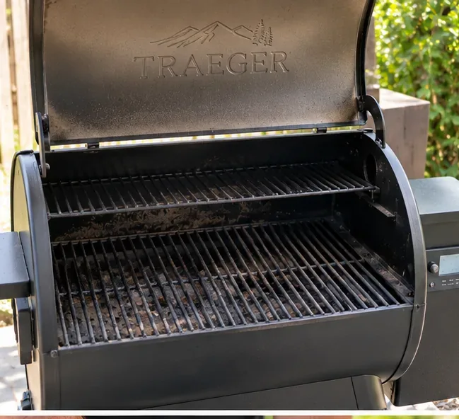 Een open Traeger pelletgrill met donkere vet- en asresten zichtbaar op de roosters en in de kookkamer.