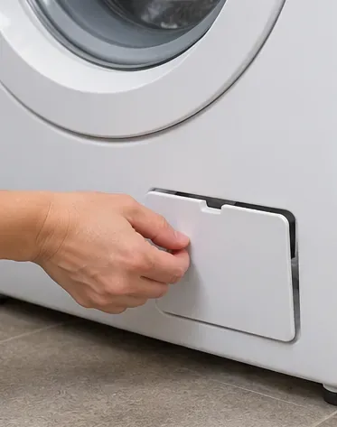 洗濯機の前面下部にあるサービスパネルを開けて、排水ポンプのフィルターにアクセスしている手。