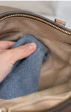 Een hand dept de vlek op de stoffen voering van een handtas met een zachte blauwe microvezeldoek.