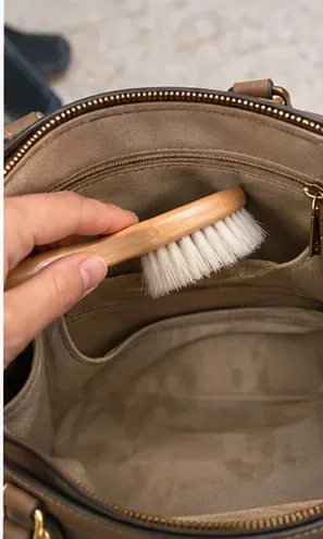 Een hand gebruikt een zachte borstel om stof los te maken in een vakje van een handtas en langs de stoffen voering.
