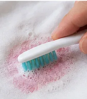 柔らかい歯ブラシを使って、白い布に残ったピンクの口紅の染みに前処理剤を優しく塗り込む。