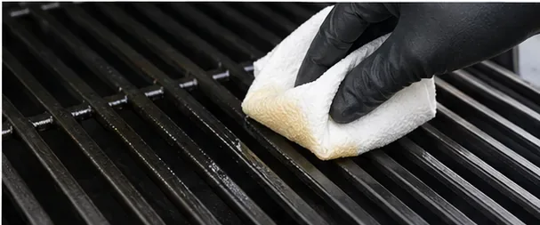 Een gehandschoende hand veegt een dun laagje bakolie over de afgekoelde grillroosters.